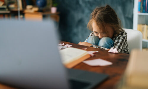 Exhausted upset primary child school girl sitting alone hugging knees in front of desk with difficult homework. Upset angry schoolgirl put head on knee being stressed with homework, selective focus.